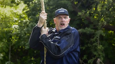 Greg Davies holding onto a rope and looking shocked at something offscreen. Trees in the background