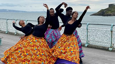 Five professional dancers perform on a pier beside a large stretch of water. Each of the dancers wears a gown from their waist covered in vibrant, colourful patterns. 