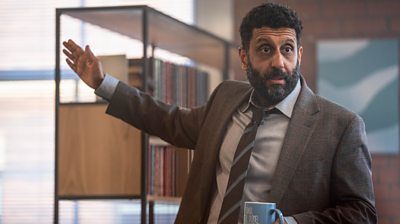 Adeel Akhtar as Sam holding a mug and his arm up looking as though he is talking to someone off screen