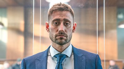 close up of Michael Socha wearing a suit and looking dejected.  He's in a glass case which implies he's in court