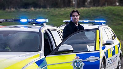 Joe Dempsie coming out of a police car and looking concerned over the door 