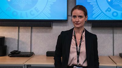 Helen McGuire looking to the camera with a police lanyard on. There are screens in the background with a police logo on them