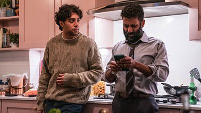 Ali Khan and Adeel Akhtar leaning on a counter in a kitchen. Adeel is looking concerned at his phone while Mani looks at him