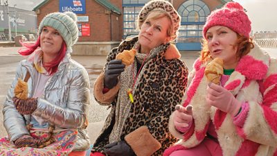 Lin (Siobhan Finneran), Joan (Lorraine Ashbourne), and Alma (Sophie Willan), sit next to each other on a bench outside an RNLI centre. Dressed in warm clothing, including wooly hats and gloves, each of the women hold a croissant in their hand, as they look at something off-screen. 