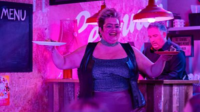 Leanne (Jayde Adams) holds two plates of food aloft as she smiles in a restaurant. Kitchen staff work in the background. A chalkboard sign on the wall beside Leanne says “Menu” but is otherwise blank. 