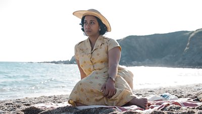 Anjana Vasan as Mary Aldin in Towards Zero. A woman sits on a blanket on a sandy beach. She wears a yellow dress covered in a flower pattern, with an accompanying yellow sunhat. The sea glistens in the sunlight in the background. 