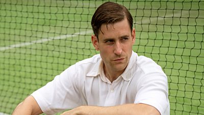 Oliver Jackson-Cohen as Nevile Strange. Close-up of a man sat leaning against a net on a tennis court. He wears a white polo neck and stares at something offscreen. 