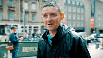 Sonny, wearing a black rain coat, walking through Edinburgh's Grassmarket area