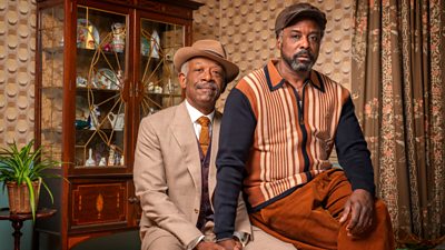 Characters Barry Walker (Lennie James) and Morris De La Roux (Ariyon Bakare). They're both sitting down holding on another hand and looking to the camera. brown and orange decor and colour scheme