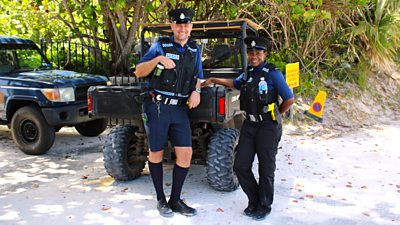 Two police offers in uniform lean against an off-road vehicle and smile to the camera. 