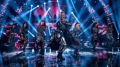 Professional dancer Johannes Radebe leads Strictly female pro dancers in a Beyonce inspired routine. They all wear long black coats and dance in formation on the ballroom floor in heels, with blue and pink lighting.