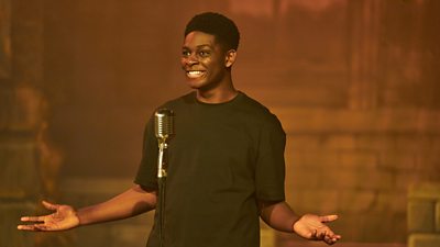 Close-up of Femi (Aruna Jalloh) standing in front of a microphone on a stage