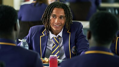 Close up of Toby (Sekou Diaby) sat in a canteen with other students wearing school uniform. 