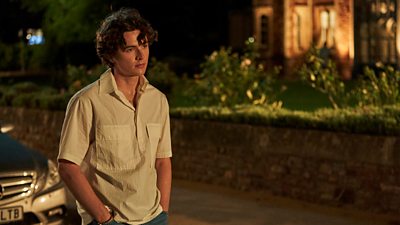 Rupert (Harry Gilby) stands on the road outside a house at nighttime. A car is parked behind him.  