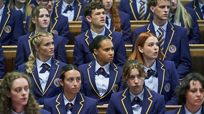 Audience of students in matching school uniforms. Florence (Rosie Graham), Abby (Assa Kanouté) and Beatrix (Tallulah Greive) sit next to each other in the center of the picture and stare at something off-screen in amazement. 