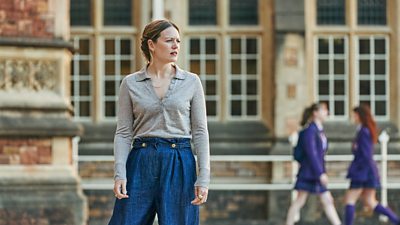 Jude (Cara Theobold) stands in the grounds of a grand building. Students in school uniform walk in the background. 