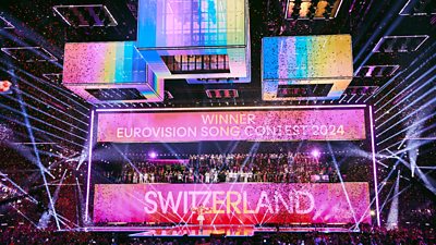 Singer Nemo stands on stage in the crowded arena as Switzerland is declared winner of the Eurovision Song Contest 2024. Confetti falls from the roof as beams of light criss cross the stage and all the Eurovision acts applaud from their backstage area, which is revealed as the main screen splits open.