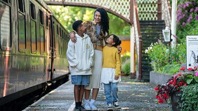 characters Perry (Tylan Bailey), Becks (Ida Brooke) And Phoebe (Ava McCarthy) walking down a train platform. Becks has her arm around the children and they're smiling