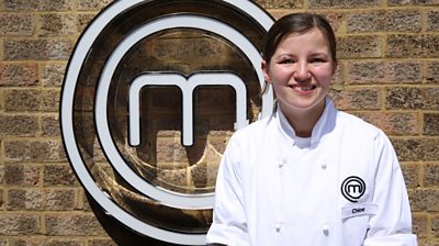 contestant Chloe stands in front of the MasterChef M, smiling at the camera and wearing a MasterChef-branded shirt