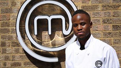 contestant Nkosi stands in front of MasterChef M looking at the camera and wearing a MasterChef branded shirt