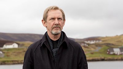 Ian Hart as Professor Euan Rossi stood outdoors beside the sea. A remote village is visible in the background behind him.