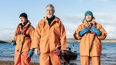 Ross Anderson, Vincent Regan and Macleod Stephen as Patrick, John and Fegrus Harris. The three characters characters stand on the beach beside a speedboat. All three wear waterproof orange clothing. 