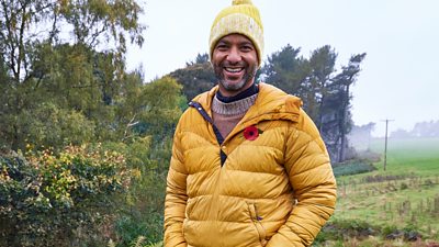 Sean Fletcher in a coat and hat smiling at the camera with grass and trees in the background