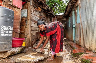 A young woman in a red printed dress and black scarf leans over broken chunks of concrete outside her home. Nearby is a wall of corrugated metal and a large water holding container