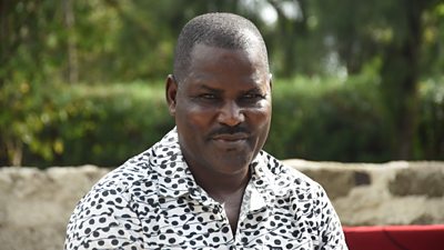 A man in a white and black patterned shirt stares at the camera