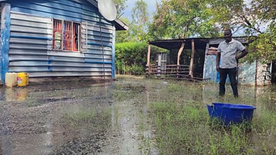 A man stands in a flooded yard with water coming halfway up his lower legs. Grass peeks through the top of the water. A shelter of corrugated steel is behind and a house, also with corrugated steel, is to the side. A blue bucket sits in the foreground,