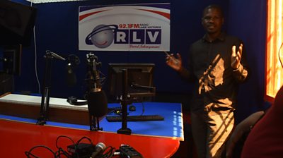 A man in dark shirt gestures as he stands in a radio studio. Behind him is a sign with RLV on the wall, and beside him is a desk with microphones and a screen.