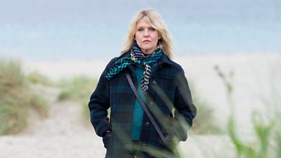 Photo of Ashley Jensen (Calder) stood amidst sand dunes beside a beach with her hands in her coat pockets.