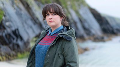 Photo of Alison O’Donnell (Tosh) stood on a beach beside a damp, moss-covered cliff. 