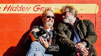 Dave Myers and Si King sitting in their biking gear leaning against an orange wall and smiling in the sun