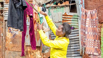 A young woman in yellow jacket hangs clothing on a line. Behind her is a rough wall of wood and corrugated metal.