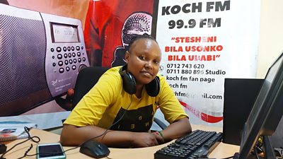 A young woman in bright yellow shirt sits behind a desk with headphones around her neck. In front is a computer screen and keyboard, and behind her a sign carries Koch FM's nae and contact details. A mural on the wall behind shows a radio and microphone.