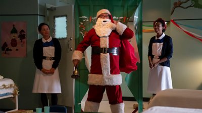 Fred Buckle (Cliff Parisi) wears a Father Christmas costume, complete with a big white beard, and a sack of presents. He stands in a hospital ward beside two characters in nurses uniforms. 