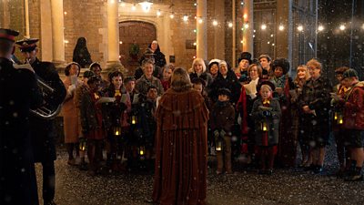 A choir of children, nurses and midwives stand outdoors in the snow. Fairylights are draped across the buildings in the background. A brass band are playing to the left of the image. Members of the choir hold song sheets and lanterns. 