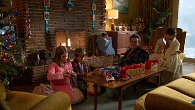 Dr Turner (Stephen McGann) sits in a living room with three children as they unwrap Christmas presents. The room is decorated with paper ribbons, a Christmas tree, and sacks full of presents. A fire is lit in the fireplace. 