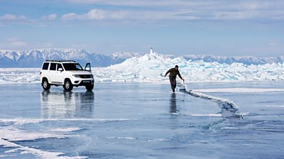 a behind the scenes shot of a crew member walking along ice with a car in the background