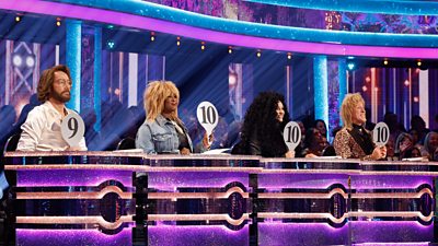 The Strictly Come Dancing judges sit behind the desks in costume for Icons Week, holding aloft their 10 panels