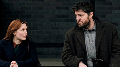 Image from Ink Black Heart showing Holliday Grainger as Robin Ellacott and Tom Burke as Cormoran Strike. The pair look at each other as they lean on a railing. 