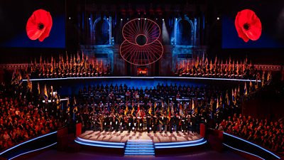 Image from the Royal British Legion Festival of Remembrance. A stage filled with people sits below large screens displaying poppies. 