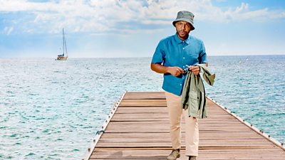 Don Gilet as DI Mervin Wilson, who walks towards the camera on a wooden jetty that stretches back into the ocean where a small boat is visible. He wears light trousers, a blue shirt and a bucket hat, holding sunglasses in his hands and a jacket over his arm.