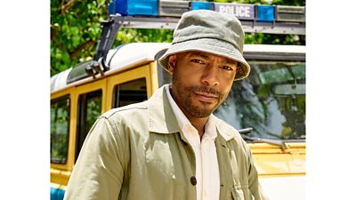 DI Mervin Wilson stands by a yellow police truck, wearing a light green jacket, white shirt and green bucket hat.