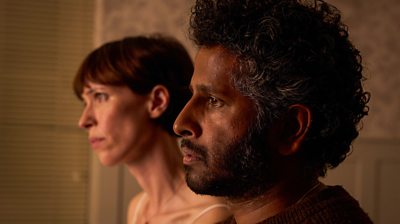 Close up of Paul Kutty (Prasanna Puwanarajah) and Claire Kutty (Rebecca Hall) behind him.  They're both looking sad and concerned facing the right 