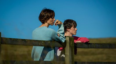 Claire Kutty (Rebecca Hall) and Kyle Francis (Ollie West) looking to the distance leaning on a fence with a clear blue sky