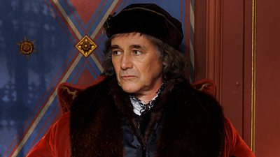 Mark Rylance in character as Thomas Cromwell. Sat in fine clothing of the Tudor period, he looks at something offscreen.