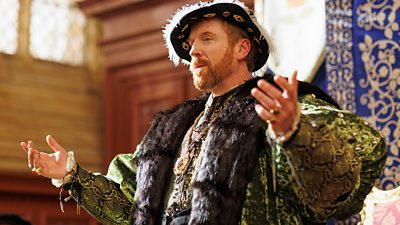 Damian Lewis in character as King Henry VIII. Standing with his arms outstretched, Henry VIII wears fine, regal clothing of the Tudor Period and stands in a grand room with high ceilings. 