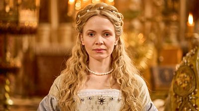 Kate Phillips in character as Jane Seymour. Dressed in fine clothing of the Tudor Period, she wears pearl jewellery and stands in a large room with lots of ornate gold furnishings. 